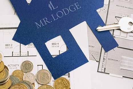 Symbolbild für Immobilieninvestitionen mit Bauplänen, Geldmünzen, Schlüssel und Mr. Lodge Logo – Ihr kompetenter Partner für möbliertes Wohnen und Kapitalanlagen in München.